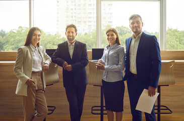Team of four successful business people at work. Group portrait of happy confident men and women in formal suits standing together by office window, looking at camera and smiling