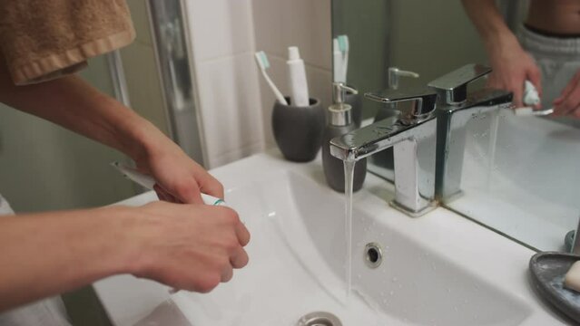 Hungover gen Z man with swollen face taking toothpaste and brushing his teeth in front of bathroom mirror in morning at home
