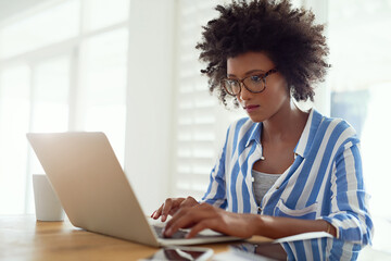 Black woman, desk and laptop in home with typing for remote work, online business and entrepreneur. African female writer, technology and research by table with book author, website and fiction story