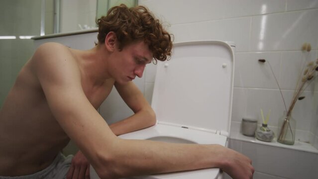 Young hungover guy leaning on toilet in bathroom while feeling nausea in morning after party at home
