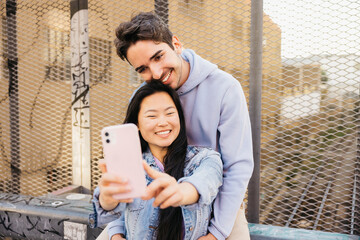 Couple sitting on a bridge over an urban railroad smiling and taking a selfie with a phone....