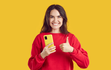 Beautiful attractive young woman holding smartphone showing thumb up. Happy girl in red sweetshirt...