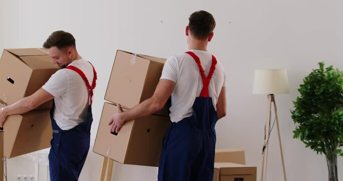 Young cheerful professional workers of moving service in overalls carrying and delivering cardboard boxes in new apartment. Move, moving day and relocation concept. 4k video. Slow motion video.