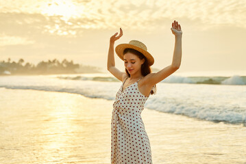 Elegant lady in polka dot dress enjoys sunset on beach with arms raised. Solo traveler feels free...