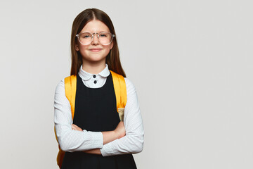 Delighted schoolgirl kid wearing uniform, eyeglasses and yellow backpack, holding crossed hands and...