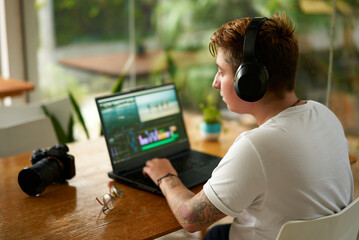 Focused individual with headphones edits on laptop inside cafe, video editing skills at work. mirrorless camera lays beside, freelance job in progress. Cafe surroundings, daylight illuminates process.