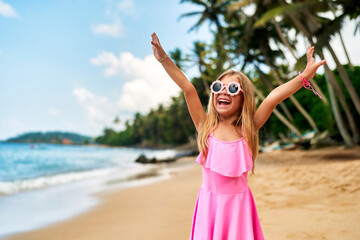 Joyful young girl in pink dress, sunglasses on sunny tropical beach with palms. Arms raised, she...