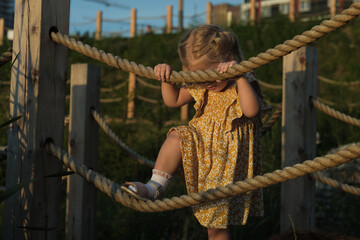 Toddler gripping rope bridge, looks on curiously. Reflects learning and exploration, a staple of...