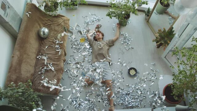 Top down view of young man tossing tinsel up in air and resting on messy floor in living room while having afterparty hangover