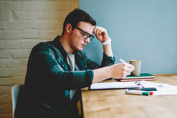 Pensive hipster guy studying in coworking space learning information from notings and textbooks for...