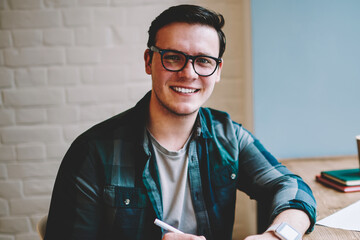 Portrait of cheerful hipster guy in spectacles enjoying studying in coworking space using wifi for...