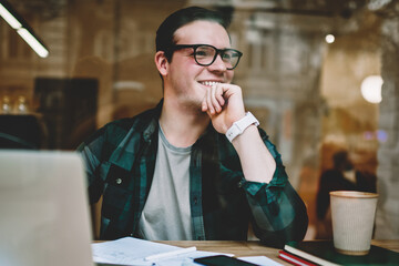 Prosperous male freelancer in good mood satisfied with completing project sitting with laptop computer in cafe, smiling hipster guy creating article for web page publication happy about great idea