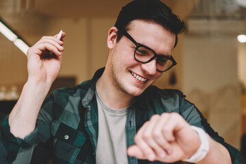 Cheerful male student satisfied with useful apps on digital smartwatch looking at touchscreen,...