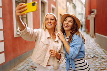 Two beautiful young woman friends walking together around the city, using smartphone while making selfie. Fashion, travel, technology, blogging concept.