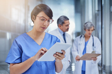 Woman, doctor and tablet in hospital with scrubs for medical, check on digital file for schedule. Female surgeon, medicine and tech with colleagues in background and working, career in healthcare