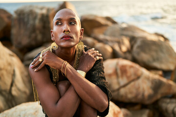 Non-binary black person in luxury dress, golden jewelry on beach rocks in ocean. Trans ethnic...