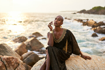 Non-binary black person in luxury dress sits on rocks in ocean. Trans ethnic fashion model wearing jewelry in posh gown, poses in tropical seaside location. Divine feminine human. Diversity concept.