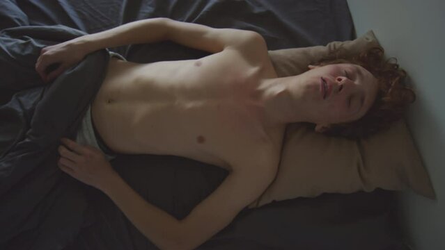 Top down view of young shirtless guy falling back in bed and sleeping while suffering from hangover in morning after party