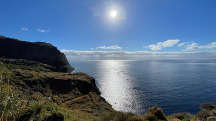 A stunning coastal landscape with sunlight reflecting off the calm ocean, set against cliffs and a clear blue sky.