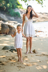 Young woman mother with a little daughter in white dresses having a fun on seashore in the shade of trees and palms