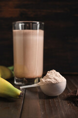 Plastic measuring spoon with whey protein powder, chocolate milkshake cocktail in a glass, blended protein drink and banana fruit on a dark wooden background