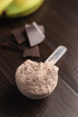Plastic measuring spoon with whey protein powder, chocolate cubes and banana fruit on a dark wooden background