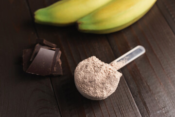 Plastic measuring spoon with whey protein powder, chocolate cubes and banana fruit on a dark wooden background