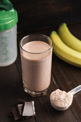 Protein milkshake cocktail in a glass, plastic measuring spoon with whey protein powder, shaker for prepare blended protein drink, chocolate cubes and banana fruit on a dark wooden background