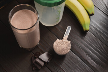 Plastic measuring spoon with whey protein powder, milkshake cocktail in a glass, shaker for prepare blended protein drink, chocolate cubes and banana fruit on a dark wooden background