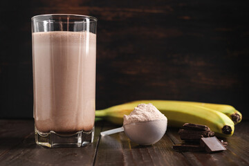 Plastic measuring spoon with whey protein powder, chocolate milkshake cocktail in a glass, blended protein drink and banana fruit on a dark wooden background