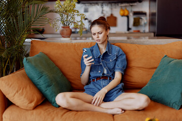 Young woman sitting on orange couch with smartphone in hand, engrossed in screen, modern technology...