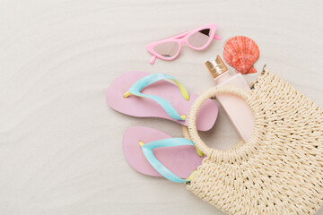 Straw bag with accessories on sand background, top view