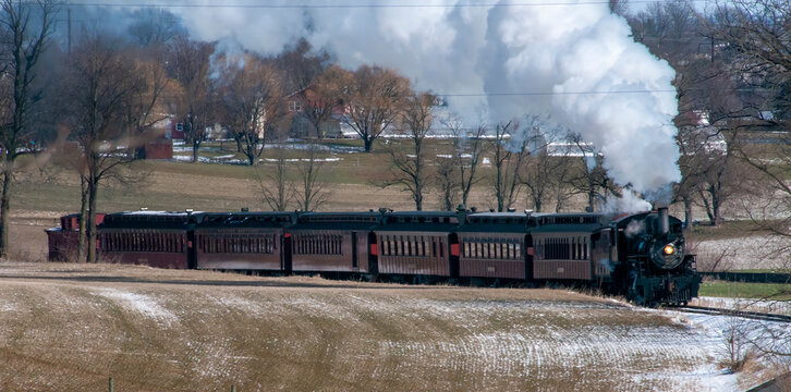 A steam train is traveling down a snowy track. The train is black and white and is surrounded by trees. The train is the main focus of the image, and the snowy landscape creates a serene