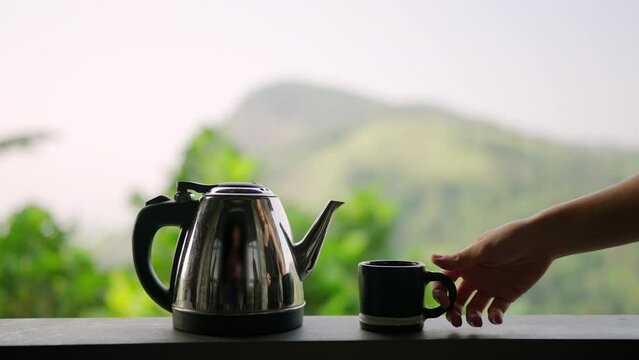 Woman enjoys fresh brewed tea on scenic terrace overlooking rich mountain, plantation. Hand reaches for cup, reflects relaxing moment in nature. Beverage preparation with tranquil background. Slowmo