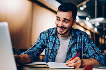 Cheerful hipster guy laughing at social networks content browsed on laptop computer during homework preparation in library, happy male student watching funny videos on netbook while learning