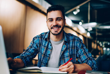 Portrait of cheerful male student enjoying learning in coworking office using laptop computer for research,happy freelancer looking at camera during making project for remote job making notes