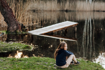 Little red-haired girl, lonely dreaming child sits on green grass against the background of a lake in nature outdoors on vacation in the countryside at sunset. Photography, portrait, landscape.