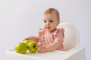 Fototapeta premium Cute baby in a baby chair nibbles apples on a white background