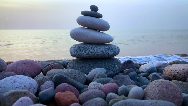 Balanced sea stones on a pebble beach with foaming and waving sea. Balanced pebble stones pyramid close up. Calm, harmony, zen, spa, relaxing meditation background. Sea water waves crashing at sunset.