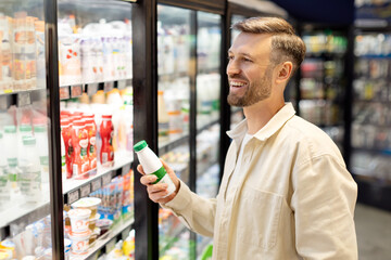 Handsome man doing shopping in supermarket, standing near fridge with dairy products, buying...