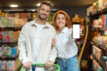 Happy spouses standing at supermarket and showing blank smartphone, shopping and recommending app...