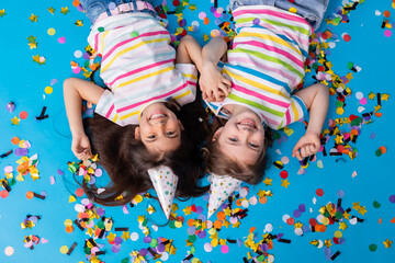 Two cute girls in birthday hats laughing, confetti, blue background, top view