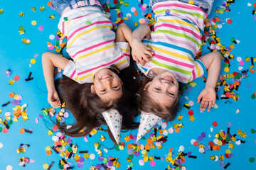 Two cute girls in birthday hats laughing, confetti, blue background, top view