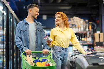 Happy young couple shopping together at supermarket, husband and wife standing by freezer with...