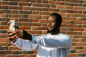 Happy young African American man in dashiki ethnic clothes taking selfie on brick wall background....