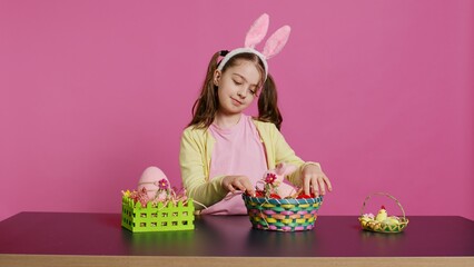 Excited young girl arranging painted eggs in a basket to prepare for easter holiday celebration,...