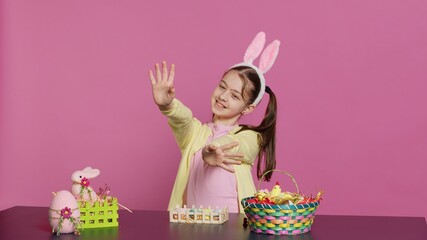 Obraz premium Energetic young girl with adorable bunny ears waving in studio, saying hello and greeting someone while she creates easter decorations. Joyful toddler posing against pink backdrop. Camera A.