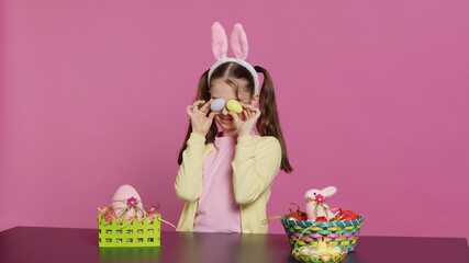 Obraz premium Enthusiastic young girl playing peek a boo in front of camera, using painted easter eggs against pink background. Joyful lovely toddler feeling excited about spring holiday festivity. Camera A.