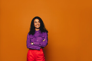 Thoughtful woman with curly hair standing on vibrant orange background, arms crossed and smiling