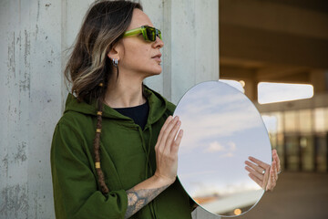 Young woman with piercings and tattoos thoughtfully holding mirror and looking into the distance